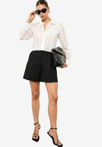 Blusa blanca con bordado floral, pantalones cortos negros de cintura alta y sandalias negras con tiras. Sostiene un bolso negro de piel acolchada y lleva gafas de sol oscuras.