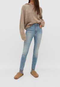 Woman wearing beige oversized knit sweater, light blue skinny jeans, gray socks, and tan slip-on clogs, standing on a plain white background.