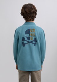 Tricou polo turcoaz cu mâneci lungi, având pe spate un grafic de craniu și oase încrucișate, cu text galben "SCALPERS CO."
