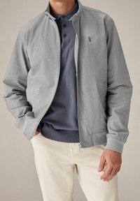 Grijze lichte bomberjack met een ritssluiting aan de voorkant, geribde manchetten en kraag, gedragen over een donkerblauw polo-shirt en crèmekleurige broek.