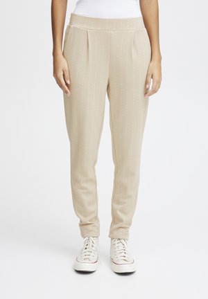 Beige tapse broeken met verticale witte streepjes, geplooide voorkant, elastische tailleband en enkel lengte, gecombineerd met witte sneakers.