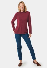 Burgunderfarbene Langarmbluse mit hohem Kragen, kombiniert mit blauen Jeans und braunen Slippern. Glatter Stoff, klassischer Schnitt.