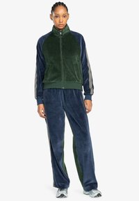Chándal de terciopelo en azul oscuro y verde, con un alto collar, cierre de cremallera y detalles a rayas en las mangas y los pantalones.