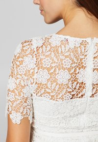 Vit spetsklänning med blommönster och korta ärmar. Den fina spetsen står i kontrast till ett inre av fast tyg, vilket visar upp intrikata detaljer.