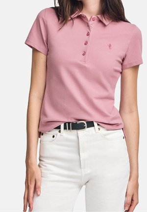 Femme portant un polo rose à manches courtes rentré dans un jean blanc taille haute avec une ceinture noire sur un fond uni.