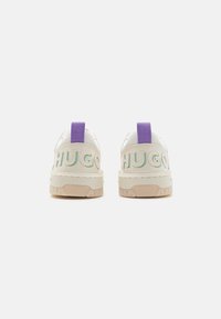 Baskets blanches avec des onglets de talon violets et un logo "HUGO" vert à l'arrière. Présente des semelles en caoutchouc texturé et un matériau supérieur lisse.