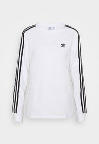 Baltas garām piedurknēm krekls ar melno Adidas logo uz krūtīm un trim melnām svītrām, kas stiepjas pa katru roku. Kokvilnas materiāls ar ribotām aprocēm.