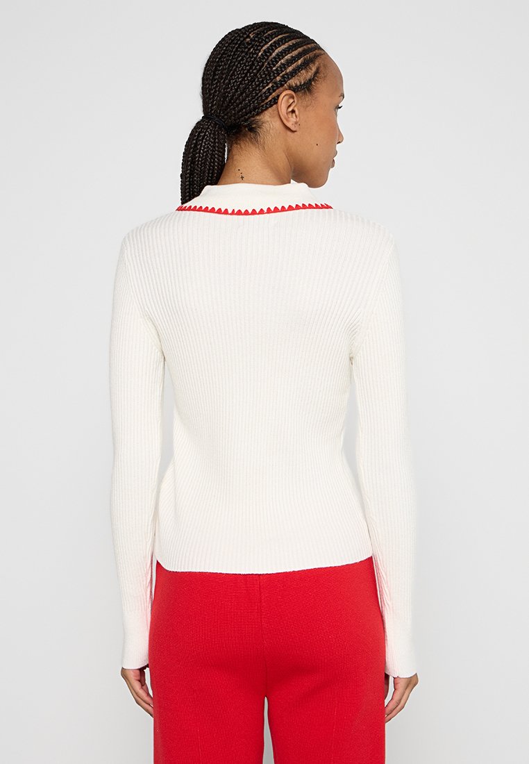 Weißer gerippter Pullover mit rotem gewellten Kragen, langen Ärmeln und taillierter Passform. Kombiniert mit roten Hosen, die einen eleganten Stil zeigen.