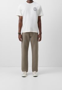 Krémové tričko s černým kruhovým logem na levé straně hrudníku, v kombinaci s béžovými kalhotami rovného střihu a bílými teniskami. Jednoduchý design, neformální vzhled.