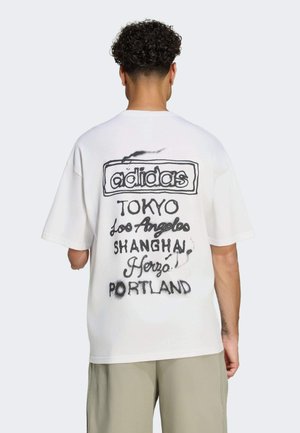 Uomo con capelli ricci che indossa una maglietta bianca Adidas con scritte nere che elencano città sul retro, in piedi di spalle alla fotocamera.