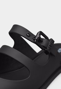 Μαύρο σανδάλι με ρυθμιζόμενο λουράκι με μαύρη αγκράφα και οπές για προσαρμογή μεγέθους, κατασκευασμένο από μαλακό ματ υλικό.