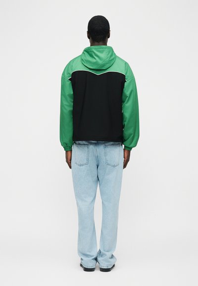 Άνδρας που στέκεται με την πλάτη προς τα εμπρός, φορώντας πράσινο και μαύρο μπουφάν με κουκούλα, ανοιχτό μπλε τζιν και μαύρα παπούτσια, σε απλό φόντο.