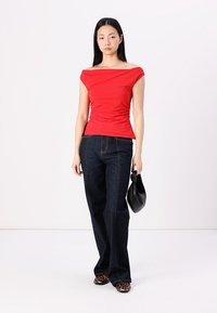 Röd off-shoulder topp med rynkdetaljer, kombinerad med mörka denim jeans med utsvängd nederdel. Accessoarer inkluderar en svart handväska. Mönstrade skor.