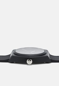 Montre noire avec un bracelet en silicone texturé et un verre circulaire lisse. Présente une couronne en argent et des éléments de design minimalistes.