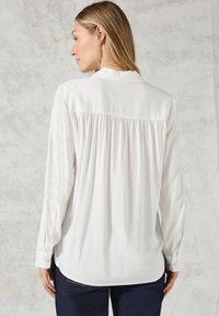 Blouse blanche à manches longues avec un détail de pli au dos, ourlet arrondi et poignets boutonnés. Texture lisse et tissu drapé doux.