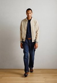 Lys beige bomberjakke med marineblå striper på mansjettene, svart t-skjorte, blå jeans og brune joggesko, båret av en person som går på tregulv.