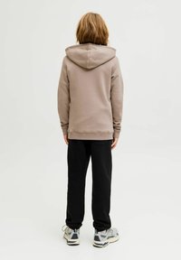 Una sudadera marrón claro con una capucha amplia, combinada con pantalones de chándal negros y zapatillas grises con detalles azules, vista desde la parte de atrás.