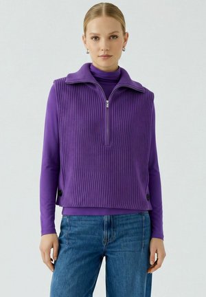 Vrouw met blond haar die een paarse gebreide mouwloze gilet draagt over een paarse lange mouwen top en blauwe spijkerbroek, staand tegen een lichte achtergrond.