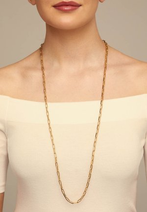 Mujer con una blusa cremosa con los hombros descubiertos y un collar largo de cadena dorada con eslabones rectangulares, sobre un fondo neutro.