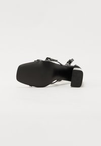 Chaussure à talon haut noire avec un talon bloc solide, texture lisse, brides de cheville ajustables et un design de bout carré large.
