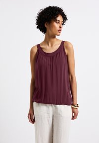 Blusa sin mangas burdeos con escote redondeado, detalle fruncido en el pecho y un corte holgado, combinada con pantalones de lino claros.
