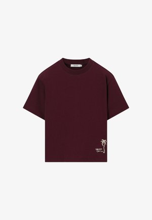T-shirt à manches courtes bordeaux foncé avec col rond, petit motif blanc de palmier et texte "Sunny spines & palm trees" près de l'ourlet.