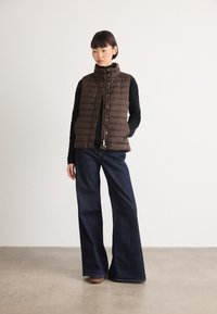 Kvinne som står, iført en brun quilted vest, svart genser, vide mørkeblå jeans og brune sko mot en ensfarget bakgrunn.