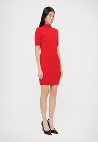 Rochie roșie tricotată, cu mâneci scurte, guler înalt și siluetă cambrată. Pantofi negri cu toc înalt completează ținuta.