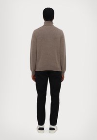 Brauner Rollkragenpullover mit langen Ärmeln, aus einem strukturierten Stoff, kombiniert mit schwarzen Hosen und weißen Sneakers mit schwarzen Akzenten.