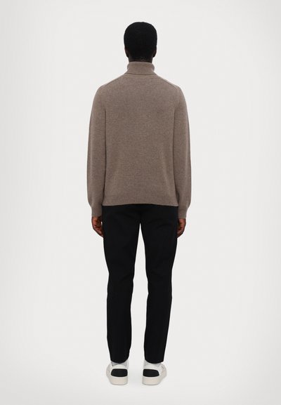 Bruine coltrui met lange mouwen, gemaakt van een gestructureerde stof, gecombineerd met zwarte broek en witte sneakers met zwarte accenten.