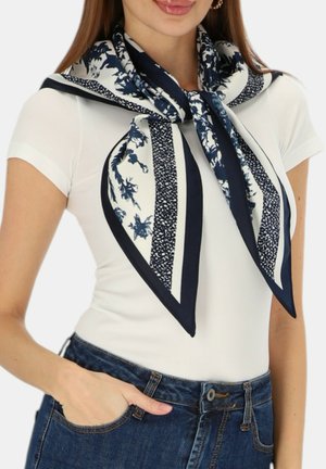 Kobieta nosząca granatowo-biały szal w kwiatowy wzór zawiązany wokół szyi, biały krótki rękaw i niebieskie dżinsy z ręką w kieszeni.