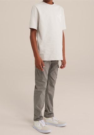 Personne portant une chemise beige clair à manches courtes, un pantalon cargo gris et des baskets blanches, debout devant un fond beige uni.