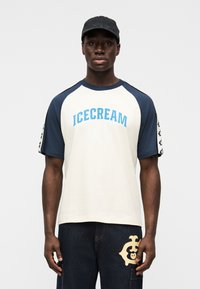 Κρεμ t-shirt με μπλε raglan μανίκια και το κείμενο "ICECREAM". Συνδυασμένο με σκούρο τζιν που διαθέτει κρεμ λογότυπο. Μαύρο καπέλο.