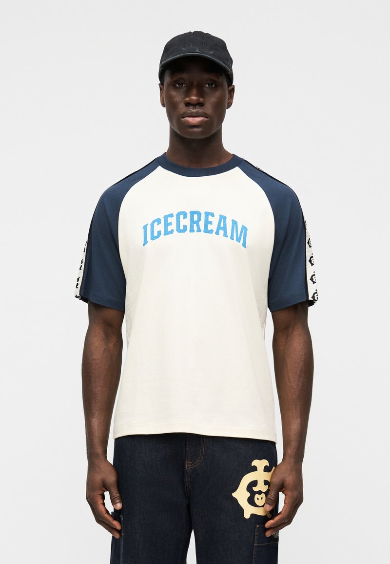 Κρεμ t-shirt με μπλε raglan μανίκια και το κείμενο "ICECREAM". Συνδυασμένο με σκούρο τζιν που διαθέτει κρεμ λογότυπο. Μαύρο καπέλο.