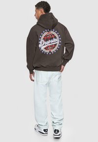 Brauner Hoodie mit einem großen, bunten Grafikdruck auf dem Rücken, der den Text "Dropsize" zeigt. Kombiniert mit hellblauen Jeans und schwarz-weißen Sneakers.