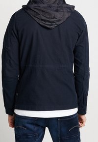 Mörkblå jacka med vadderad huva, med en reguljär passform, texturerad tyg och kontrasterande detaljer vid axlarna och ärmarna.