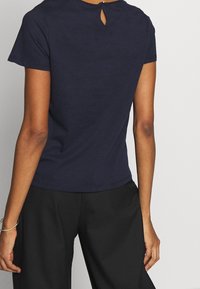 Námořnická modrá tričko s krátkým rukávem a detailem klíčové dírky na zadní straně krku. Měkká, hladká látka; jednoduchý a přiléhavý design.