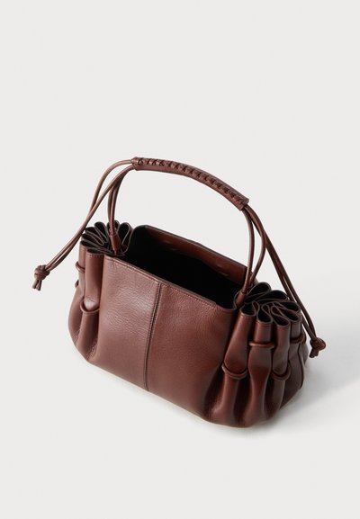 Καφέ δερμάτινη τσάντα με σφιχτές πλευρές, ενιαίο πλεγμένο χερούλι και ανοιχτό πάνω μέρος σε απλό φωτεινό φόντο.