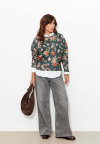 Pull vert à motif floral avec poignets côtelés, associé à une chemise blanche à col et un pantalon large en denim gris. Tenant un sac marron.