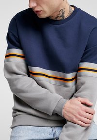 Felpa navy e grigia con righe orizzontali arancioni e grigie. Presenta un colletto a girocollo e polsini a costine. Texture morbida e design casual.