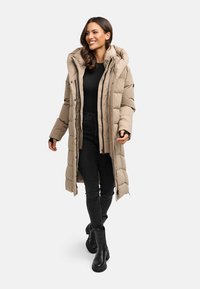 Piumino beige con colletto alto e cappuccio, dotato di chiusura con zip e bottoni a pressione, abbinato a jeans neri slim-fit e stivaletti neri.