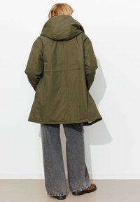 Veste à capuche vert olive à coupe décontractée, avec une texture lisse et une taille cintrée, associée à un pantalon gris évasé et des chaussures à motifs.