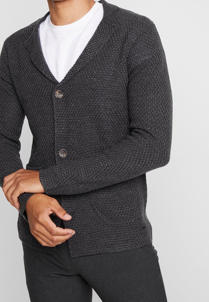 Homme portant un cardigan tricoté gris foncé texturé avec deux boutons sur un t-shirt blanc, bras croisés devant, sur fond uni.