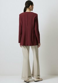 Burgundski blazer z oblikovanimi rameni in razporkom na hrbtu, v kombinaciji z kremnimi širokimi hlačami in bež loaferji, prikazan od zadaj.