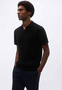 Μαύρο πλεκτό πόλο με κάθετες λεπτομέρειες, κοντά μανίκια και γιακά σε σχήμα V, συνδυασμένο με μπλε ναυτικό παντελόνι, πάνω σε απλό φόντο.