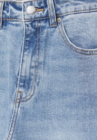 Gros plan sur le devant d'un jean en denim bleu clair avec bouton en métal, passants de ceinture et poche incurvée bordée par une couture orange.