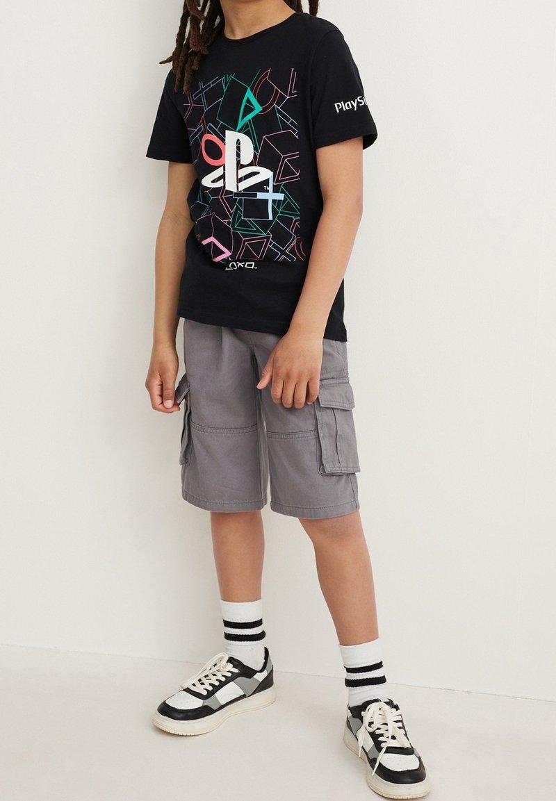 Enfant portant un T-shirt PlayStation noir, un short cargo gris, des chaussettes blanches avec des rayures noires, et des baskets noires et blanches, debout contre un mur uni.