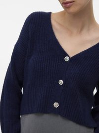 Kvinna som bär en marinblå ribbad stickad cardigan med tre dekorativa runda kristallknappar över ett grått plagg.