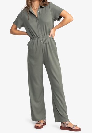 Femme portant une combinaison vert olive à manches courtes avec boutons et poches latérales, et des sandales compensées à motif floral, debout les mains sur les hanches.