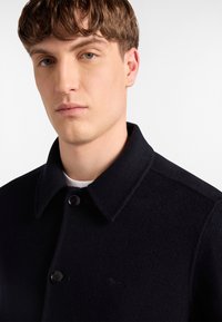 Cappotto in misto lana nero con colletto strutturato, due bottoni e un sottile dettaglio del logo. Tessuto con una superficie liscia e una vestibilità sagomata.